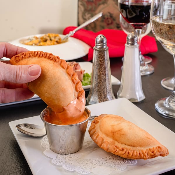 A hand dips a golden-brown empanada, labeled "CRAVINGS," into a small cup of orange sauce.