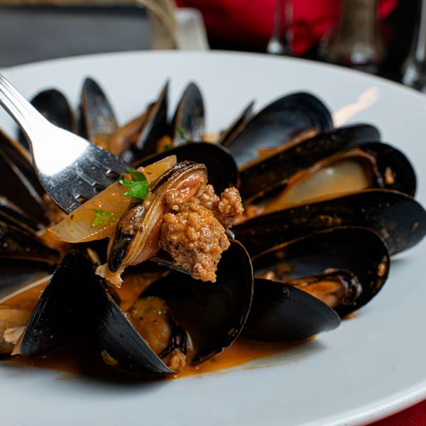 A fork lifts a mussel and sausage from a white bowl of mussels in a rich sauce.