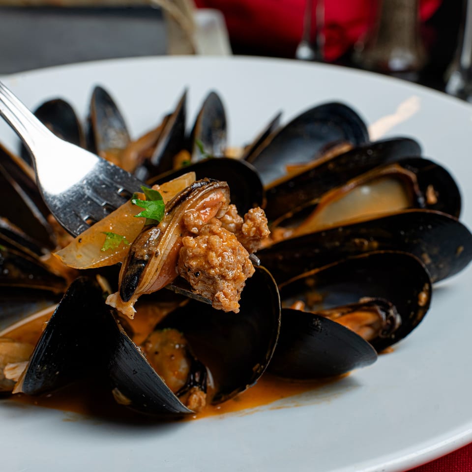 A fork lifts a mussel and sausage from a white bowl of mussels in a rich sauce.