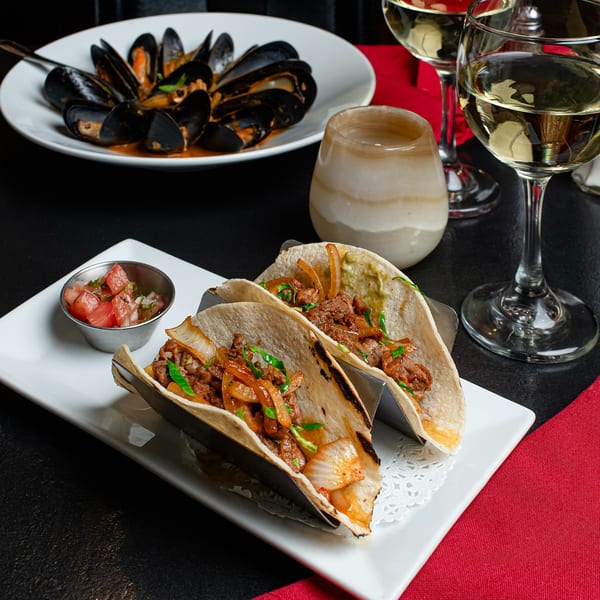 Steak tacos with onions and salsa, mussels, and white wine on a black and red table.