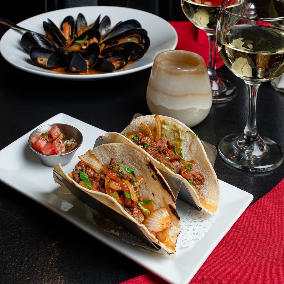 Steak tacos with onions and salsa, mussels, and white wine on a black and red table.