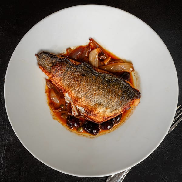 Pan-seared fish with crispy skin, served on a white plate with onions, olives, and capers in sauce.