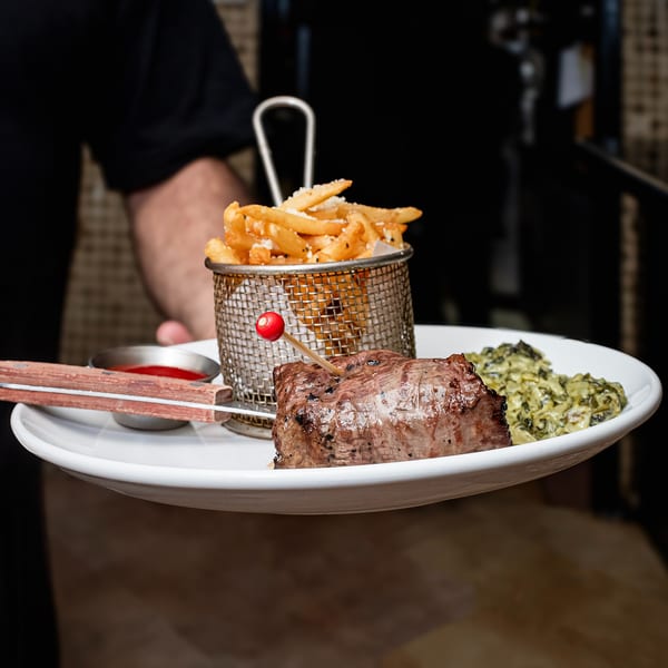 Waiter holding a plate with steak, fries in a metal basket, creamed spinach, and ketchup.