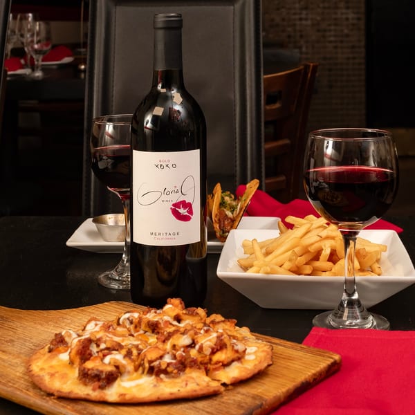 Wine bottle and glasses with flatbread, fries, and tacos on a dark table.